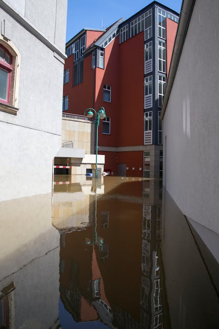 Photo of a flood-affected urban area showing a narrow alley between modern multi-storey buildings with large glass windows and red facades, partially submerged in muddy water. The water level is appro