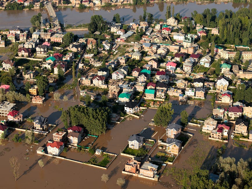 Photo of a flood-affected urban area showing a narrow alley between modern multi-storey buildings with large glass windows and red facades, partially submerged in muddy water. The water level is appro