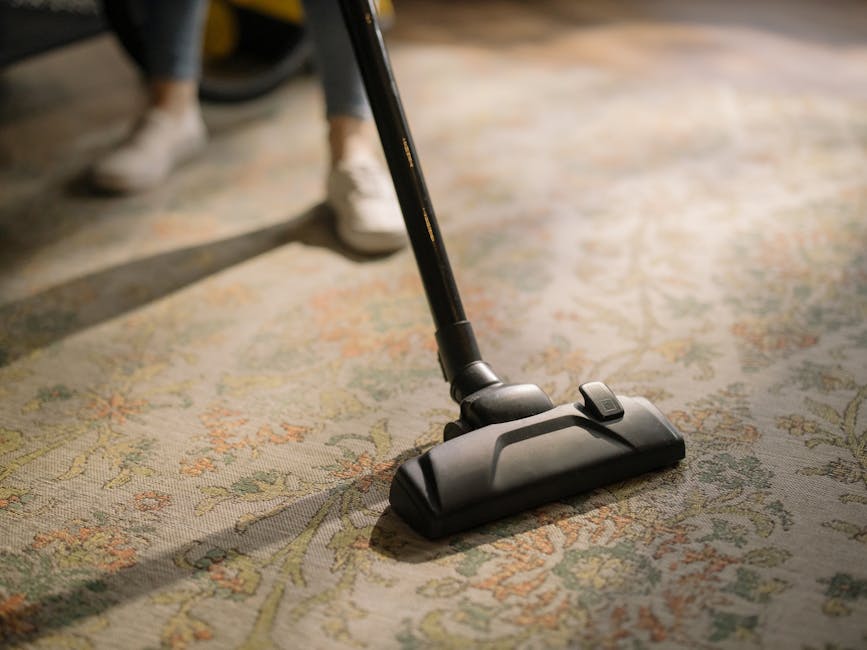 A person operating a yellow vacuum cleaner to perform deep carpet cleaning and surface sanitisation in a living room. The carpet features an intricate floral pattern in muted tones, and the cleaning p